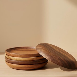 May include: A stack of five wooden plates in varying shades of brown, with one plate slightly tilted to the side. The plates have a smooth, natural wood grain finish, suggesting a rustic or minimalist aesthetic.