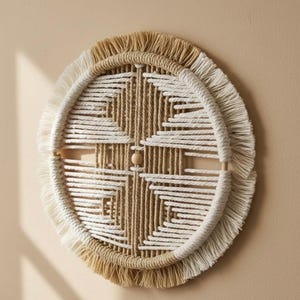 Könnte beinhalten: Ein runder, gewebter Wandbehang mit geometrischem Design. Das Stück hat eine natürliche beige-weiße Farbpalette mit einem Fransenrand. Eine kleine Holzperle befindet sich in der Mitte des Designs. Die Wanddekoration ist an einer neutralfarbenen Wand befestigt.