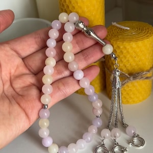 Peut inclure: Un chapelet de perles avec des perles roses pâles, lavande et crème. Les perles sont enfilées avec un connecteur et un pompon en métal argenté. Deux bougies jaunes sont en arrière-plan.