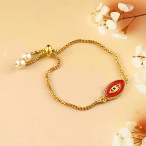Könnte beinhalten: Ein goldfarbenes Box-Kettenarmband mit einem zentralen roten Emaille-Augenanhänger mit schwarzer und weißer Pupille. Das Armband verfügt über einen verstellbaren Schiebeverschluss, der mit zwei kleinen weißen, kugelförmigen Perlen an den Enden versehen ist.