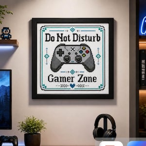 May include: A square framed cross-stitch wall art piece featuring a grey video game controller graphic centred between the text Do Not Disturb and Gamer Zone. The design includes teal geometric accents and a small heart, all on a white background.