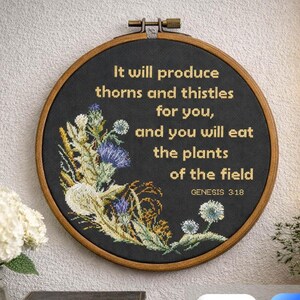May include: A round, black cross-stitch embroidery hoop with a botanical design and text. The text reads "It will produce thorns and thistles for you, and you will eat the plants of the field GENESIS 3:18." The embroidery features blue and white flowers.