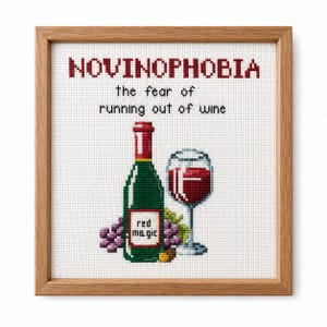 May include: A framed cross-stitch embroidery featuring the text NOVINOPHOBIA the fear of running out of wine. The design includes a green wine bottle labelled red magic, a glass of red wine, and purple grapes, all set against a white fabric background.