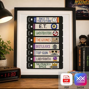 May include: A framed cross-stitch pattern featuring a vertical stack of eight black VHS tape labels. Each label displays a movie title and a corresponding icon, including Back to the Future, E.T., Ghostbusters, The Goonies, Beetlejuice, Top Gun, Labyrinth, and The Princess Bride.