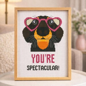May include: Cross-stitch art in a wooden frame. The artwork features a black and brown animal face wearing pink heart-shaped glasses. The text "YOU'RE SPECTACULAR!" is displayed in pink and black font.