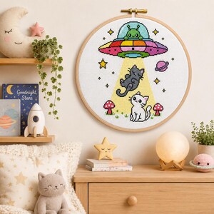 May include: A circular embroidery hoop containing a cross-stitch pattern of a green alien in a colorful UFO beaming up a gray cat. Below, a white cat sits near two red mushrooms. The design includes yellow stars and a small pink planet on white fabric.