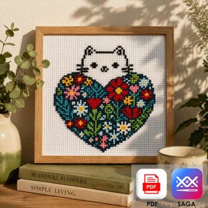 May include: Cross-stitch artwork of a white cat above a heart filled with flowers in various colours. The heart is set against a teal background and framed in a light brown wooden frame. The artwork is displayed on a shelf with books and a vase.