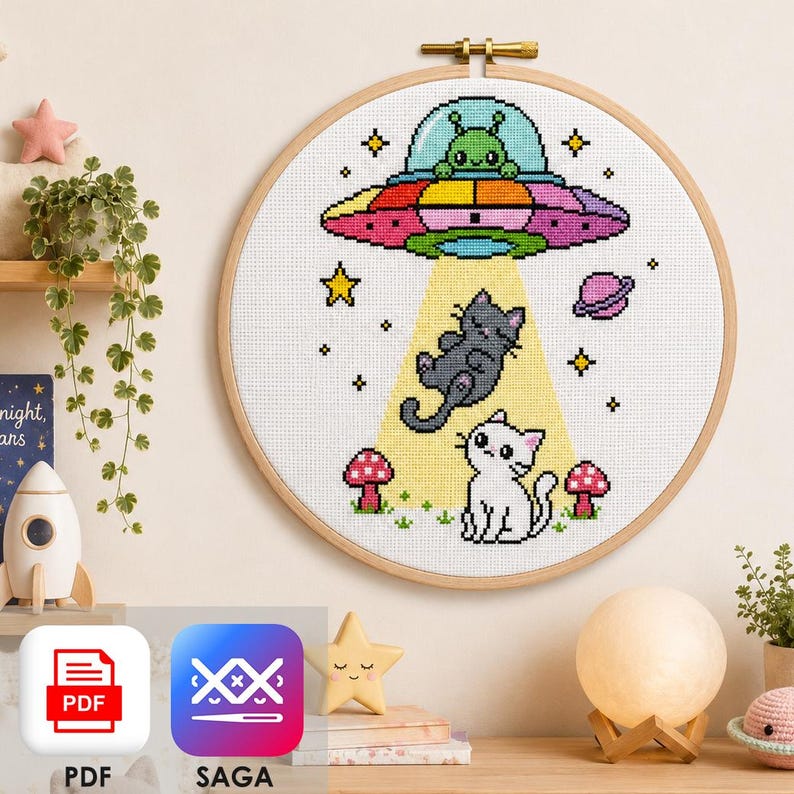 May include: A circular wooden embroidery hoop containing a cross-stitch pattern of a colorful UFO with a green alien pilot, beaming down a yellow light onto a gray cat and a white cat, surrounded by small red mushrooms and stars on white fabric.