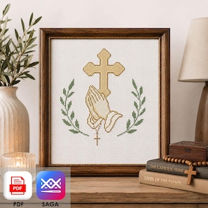 May include: A framed cross-stitch pattern featuring a gold cross above a pair of praying hands, flanked by two green leafy branches. A small cross hangs from the hands. The design is stitched on white Aida cloth and enclosed in a dark wood frame.
