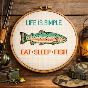 Puede incluir: Un bastidor de punto de cruz con el texto "LIFE IS SIMPLE" sobre una ilustración de un pez colorido y las palabras "EAT • SLEEP • FISH" debajo. El bastidor está sobre un fondo de madera, con aparejos de pesca y una linterna.