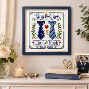 Op de afbeelding: Een ingelijst kruissteek borduurwerk met de tekst Tying the Knot, de namen Liam & Noah en de datum 06.14.2026. Het ontwerp bevat twee dassen, een rood hart, twee gouden trouwringen en groene florale accenten op een witte ondergrond.