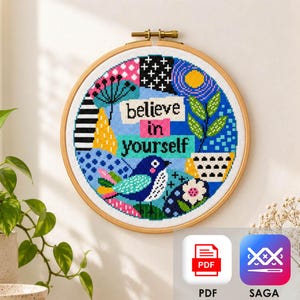 以下が含まれることがあります： believe in yourselfという文字が刺繍された、木製の円形刺繍枠。白い布地に、青い鳥、鉢植え、そしてピンク、黄色、黒、青の様々な幾何学模様がクロスステッチで表現されています。