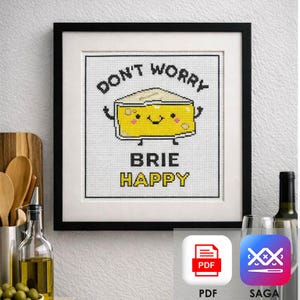 Puede incluir: Una obra de arte en punto de cruz enmarcada en un marco negro. La obra presenta un queso brie de dibujos animados sonriente con el texto "DON'T WORRY BRIE HAPPY" en negro y amarillo. La imagen se muestra en un entorno de cocina con una tabla de cortar y una botella de aceite de oliva.