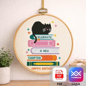 Può includere: Un motivo a punto croce in un telaio rotondo di legno raffigurante un gatto nero seduto su una pila di libri colorati. I libri riportano le scritte CELEBRATE, A NEW, CHAPTER e HAPPY BIRTHDAY, decorate con piccole stelle colorate.