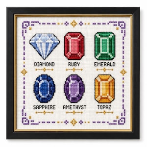 May include: A framed cross-stitch sampler on white fabric featuring six colorful gemstone illustrations: a blue diamond, red ruby, green emerald, blue sapphire, purple amethyst, and yellow topaz, each labeled with its name in black text.