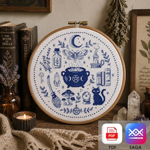 Può includere: Un ricamo a punto croce rotondo, blu e bianco, con simboli da strega. Il disegno include una luna crescente, un calderone, una farfalla e un gatto, all'interno di un telaio di legno. Il diametro è di circa 20 cm.