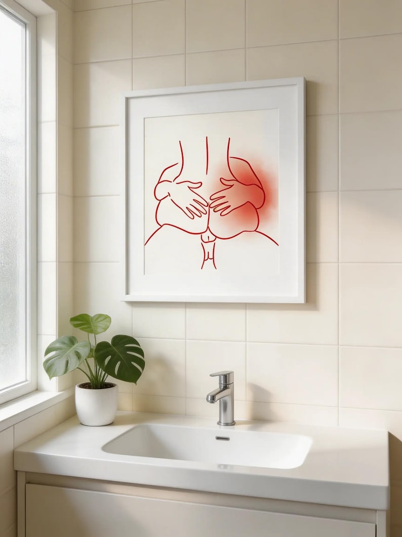 May include: A framed art print featuring a minimalist line drawing in red of a person's back and hands. The artwork is displayed in a white frame against a white background. The image is in a bathroom setting with a sink and a potted plant.