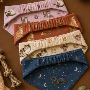 May include: A set of four triangular pet bandanas featuring elastic neckbands. The designs include a light purple cat print, a rust-colored fish bone pattern, a cream-colored cat and moon motif, and a dark blue celestial print with a black cat.