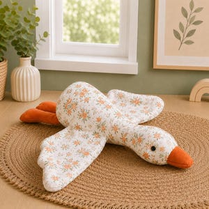 May include: A plush goose toy featuring a white fabric body covered in a small orange and green floral pattern. The goose has a solid orange beak and orange feet, with a single black bead eye visible on the side of its head. The wings are spread out.