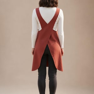 May include: A rust-colored apron with a cross-back design. The apron is worn over a white long-sleeved shirt and black pants. The model is wearing white sneakers. The apron is a kitchen or craft accessory.