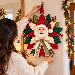 Könnte beinhalten: Ein gesteppter Weihnachtskranz aus Stoff mit einem zentralen Weihnachtsmann-Gesicht mit roter Mütze, umgeben von geschichteten Blütenblättern in Rot, Creme und Dunkelgrün. Das Design enthält karierte Stoffakzente und eine große cremefarbene Satinschleife.
