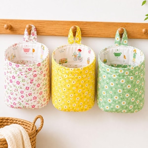 May include: Three cylindrical fabric hanging storage baskets with floral patterns in pink, yellow, and green. Each basket features a matching fabric loop for hanging and an interior lining decorated with small colorful illustrations of people and objects.