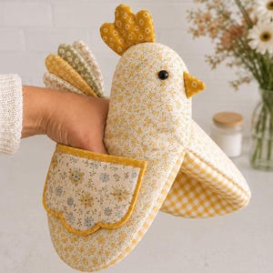 May include: A chicken-shaped oven mitt featuring a yellow and white floral print fabric with a solid yellow comb and a small yellow beak. The mitt includes a scalloped pocket with a blue and yellow floral pattern and a yellow and white gingham lining.