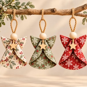 May include: Three fabric angel ornaments hanging from a branch. Each features a wooden bead head, a small wooden star accent, and a twisted jute hanging loop. The left angel is cream with red berries, the center is sage green with white snowflakes, and the right is red with white snowflakes.
