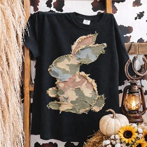 May include: Black t-shirt featuring a patchwork bunny design in camouflage colors. The bunny is made of various fabric pieces in shades of green, brown, and blue. The shirt is displayed on a wooden rack with decorative elements.