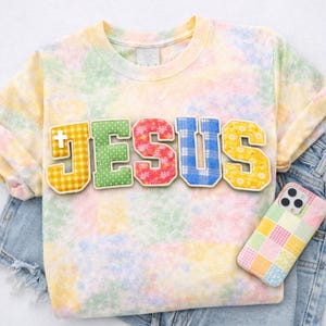 Puede incluir: Una sudadera teñida con tinte anudado en colores pastel con la palabra "JESUS" en letras aplicadas. Las letras tienen diferentes patrones: cuadros amarillos, lunares verdes, flores rosas, cuadros azules y flores amarillas. Una funda de teléfono patchwork es visible sobre unos pantalones cortos vaqueros.