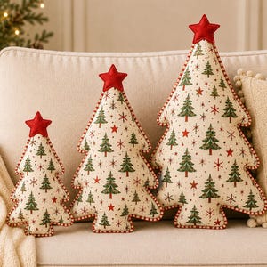 May include: Three plush Christmas tree-shaped pillows in varying sizes, featuring a cream-colored fabric with a repeating pattern of green pine trees, red stars, and snowflakes. Each pillow is topped with a red felt star and red-striped trim.