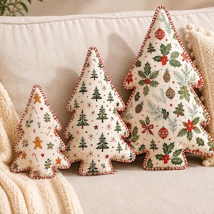 May include: Three tree-shaped decorative pillows in varying sizes, featuring off-white fabric with festive holiday prints. The smallest pillow displays gingerbread men and candy canes, while the larger two feature evergreen trees, holly, pinecones, and ornaments, all finished with red and white striped piping.
