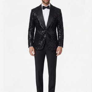 Può includere: Una giacca da smoking nera con paillettes e revers a scialle in raso, abbinata a una camicia bianca, un papillon nero, pantaloni neri sartoriali e scarpe eleganti nere lucide. La giacca presenta una texture densa e scintillante.