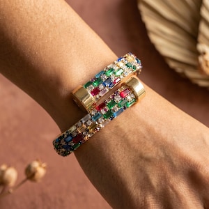 Può includere: Due braccialetti dorati con cerniera e gemme rettangolari colorate. Le gemme sono disposte a mosaico, con pietre rosse, verdi, blu e trasparenti. I braccialetti sono esposti su un polso, mostrando il design e il colore dei gioielli.