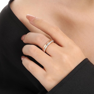 Puede incluir: Anillo de oro rosa con una banda de piedras claras y brillantes. El anillo está en un dedo, mostrando su diseño y color. El diseño del anillo es una versión moderna de un estilo clásico, adecuado para el uso diario.