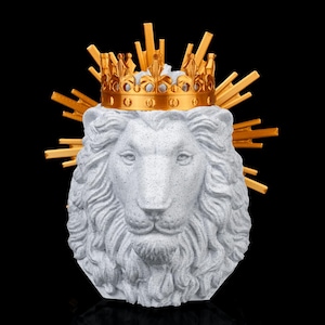 Könnte beinhalten: Eine Skulptur eines gesprenkelten hellgrauen Löwenkopfes mit detaillierter Mähne und Gesichtszügen. Der Löwe trägt eine goldene Metallkrone mit einem strahlenförmigen Design, das hinter dem Kopf hervortritt und eine strukturierte Optik erzeugt.
