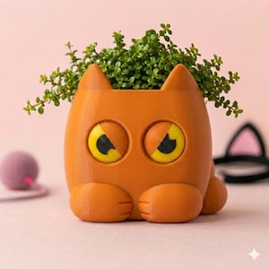 Peut inclure: Un pot de fleurs en forme de chat orange avec de grands yeux jaunes et des pupilles noires. Le pot présente deux pattes avant reposant à la base et contient une petite plante verte feuillue poussant par l'ouverture supérieure.