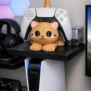 以下が含まれることがあります： ピンクの耳と黒い目の茶色の猫の置物が、白いゲームコンソールの上の黒い棚に置かれています。白いゲームコントローラーと小さな箱も棚にあります。コンソールは黒い中央部と青いライトを備えています。