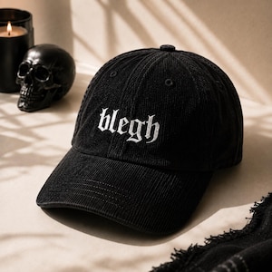 以下が含まれることがあります： フロントパネルに白いゴシック体の文字でbleghと刺繍された黒いコーデュロイ素材の野球帽。この帽子は構造化されたクラウン、カーブしたつば、そして通気のためのステッチが施されたアイレットを上部のパネルに備えています。