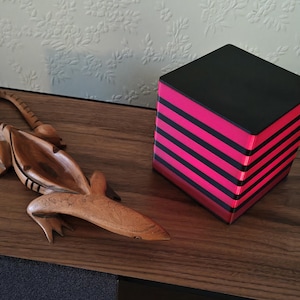 Peut inclure: Une figurine d'alligator sculptée en bois et une lampe cubique à rayures noires et roses sont posées sur une surface en bois. L'alligator est brun avec des détails sculptés. La lampe a un dessus noir et des rayures roses et noires alternées.