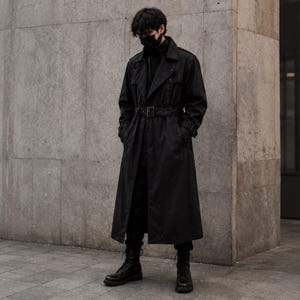 Könnte beinhalten: Ein langer schwarzer Trenchcoat mit einem Gürtel in der Taille mit rechteckiger Schnalle, einem Kragen und langen Ärmeln. Der Mantel wird über einer schwarzen Hose und schwarzen Schnürstiefeln getragen. Eine schwarze Maske bedeckt das Gesicht.