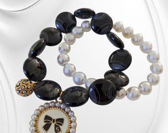 May include: Two bracelets, one with black and white patterned beads and the other with white pearls. The black and white bracelet has a gold charm with a black bow and the number "5" on it. The charm is attached to a small gold ball with a black and white eye design.