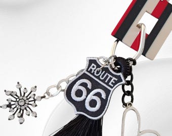 May include: A Route 66 keychain with a black and white embroidered patch, a black tassel, and a silver heart charm. A silver snowflake charm is also attached. The keychain has a red, black, and white striped accent.