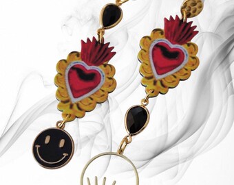 May include: Gold-coloured drop earrings with a red heart design and yellow accents, black teardrop gems, and a black smiley face charm. One earring has an eye-shaped charm. The earrings are on a white background.
