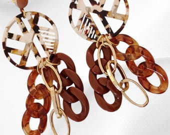 May include: A pair of statement earrings. The earrings have a circular design with a tortoiseshell pattern in shades of brown and cream. They feature a chain of brown links and gold-coloured accents, creating a bold accessory.