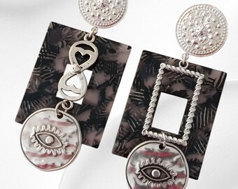 May include: A pair of silver-toned earrings. Each earring features a circular top with a floral design, a patterned rectangular pendant, and a circular charm with an eye design. One earring has a heart-shaped accent, the other a square.