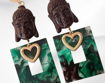 May include: A pair of dangle earrings featuring dark brown Buddha head pendants, gold-colored heart charms, and rectangular green and black marbled accents. The earrings have gold-colored oval studs.
