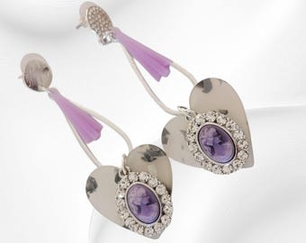 May include: Silver earrings with heart-shaped pendants. Each earring has a lavender cameo surrounded by clear crystals and a lavender accent. The earrings have a post-back closure.