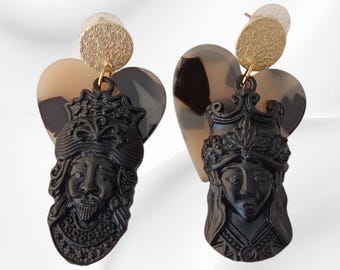 May include: A pair of black resin earrings featuring a detailed face design with a crown. The earrings have a heart-shaped backing in shades of brown and gold, with a textured gold disc at the top.