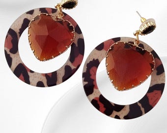 May include: A pair of dangle earrings featuring a heart-shaped, faceted, amber-colored gem, framed in gold. The gem hangs from a gold-colored post with a small black accent. The earrings are attached to a circular hoop with a leopard print design.